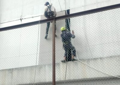 Two rope access technicians in harnesses descending a precast wall face, chain-link safety net visible around them