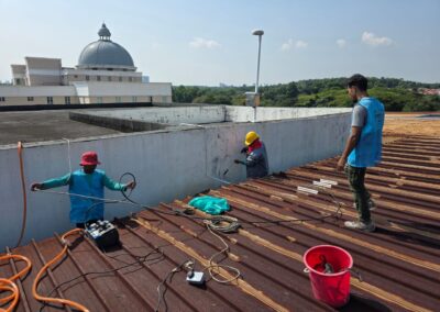 Wide rooftop view of a three-person crew preparing a concrete gutter channel with equipment laid out, school campus buildings visible in the background